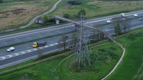 Vehicles-on-M62-motorway-passing-pylon-tower-on-countryside-farmland-fields-aerial-view-rotating-right-shot