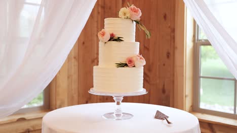 Runde,-Dreischichtige-Hochzeitstorte-Auf-Einem-Tisch-Mit-Weißem-Zuckerguss-Und-Rosa-Blumen-Und-Tortenheber-1080p-60fps