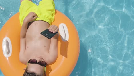 Caucasian-Boy-wearing-sunglasses-using-Smartphone-on-Orange-floater-in-the-Swimming-pool,-Top-down-view