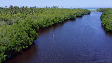 Aerial-view-of-small-boat-cruising-on-idyllic-river-surrounded-by-mangrove-forest-during-summer---Tropical-Caribbean-Sea-in-background