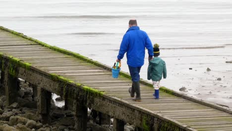 Father-holding-seaside-bucket-exploring-low-tide-UK-beach-boardwalk-jetty-with-his-son