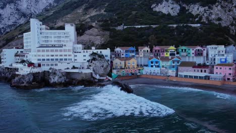 Aerial-View-Of-Waves-Breaking-At-Catalan-Bay-With-Colourful-Buildings-On-Beachfront