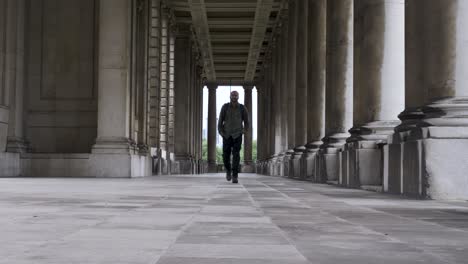 Male-Tourist-With-Backpack-Along-Colonnade-At-Royal-Navel-College-In-Greenwich-Towards-Camera