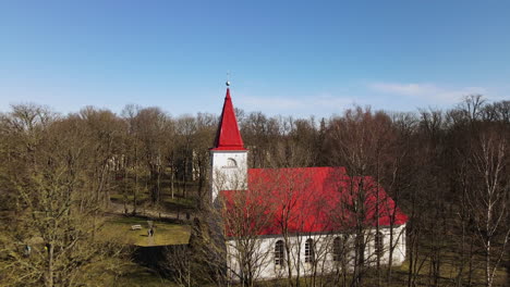Aerial-view-of-Lielvarde-Lutheran-church-at-the-bank-of-Daugava-river,-white-church-with-red-roof-through-the-leafless-trees,-sunny-spring-day,-wide-shot-moving-forward