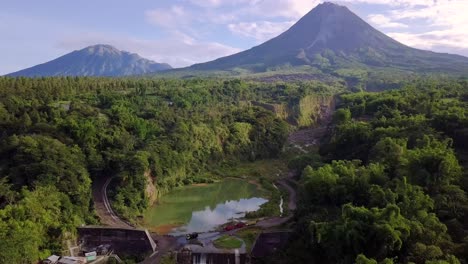 Aerial-backwards-flight-showing-natural-lake-surrounded-by-wilderness-of-Indonesia-and-giant-Merapi-Volcano-in-background-during-summer---Bego-Pendem,Indonesia