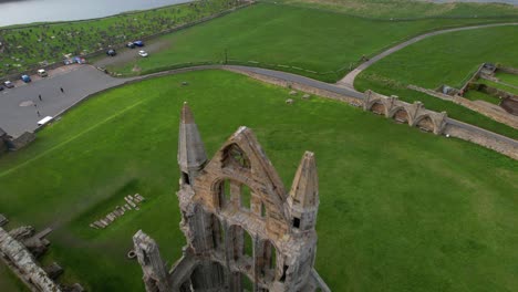 Aerial-backward-tilt-up-reveal-over-Whitby-Abbey-ruins-with-port-and-lighthouse-in-background,-North-Yorkshire-in-England