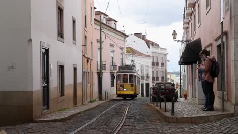 Die-Straßenbahn-28-In-Lissabon,-Portugal,-Kommt-An-Einer-Haltestelle-An,-Nachdem-Sie-Eine-Schmale,-Von-Bunten-Gebäuden-Gesäumte-Straße-Mit-Kopfsteinpflaster-Hinuntergefahren-Ist