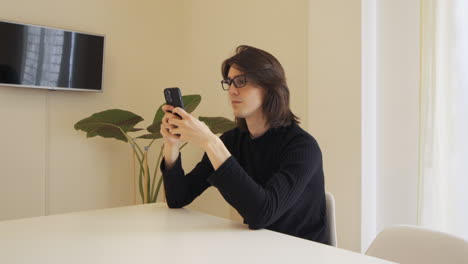 Young-man-with-glasses-in-a-beige-room-typing-on-his-cell-phone,-sitting-at-a-table