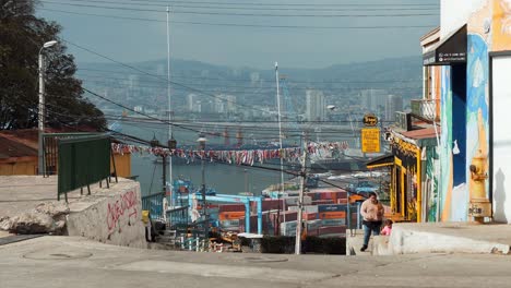 People-walking-in-uphill-street-with-picturesque-shops,-Valparaiso-Sea-Port-and-hillside-city-in-background,-Chile