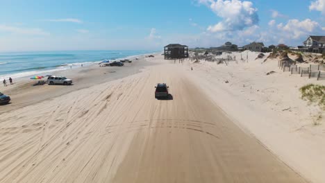Drone-following-black-suv-on-Outer-Banks-Corolla-4x4-beach,-houses-can-be-seen-inland