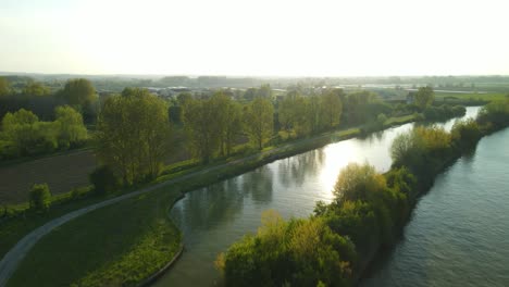 Aerial-view-in-the-sunset-going-over-a-river-revealing-the-village-of-Clairmarais,-France-in-the-distance