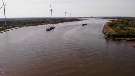 Aerial-View-Over-Oude-Maas-With-Two-Ships-Navigating-Past-Still-Wind-Turbine-On-Riverside-Bank