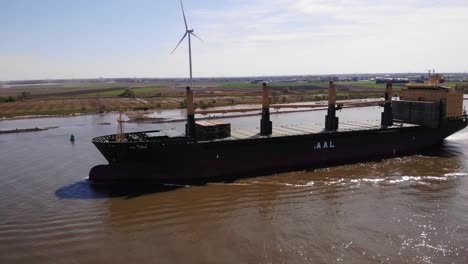 Aerial-Port-Side-View-Of-Aal-Paris-Cargo-Ship-Travelling-Along-Oude-Maas-With-Still-Wind-Turbine-In-Background-On-Sunny-Day