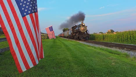 A-View-of-a-Line-of-Gently-Waving-American-Flags-on-a-Fence-by-Farmlands-as-a-Steam-Passenger-Train-Blowing-Smoke-Approaches-During-the-Golden-Hour