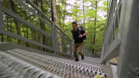 Male-hiker-with-heavy-backpack-quickly-ascends-steep-metal-staircase-in-lush-green-forest-landscape