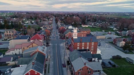 A-Drone-Approaching-View-of-a-Small-Town-and-a-Steeple-at-Sunrise-as-it-gets-Ready-to-Break-the-Horizon,-with-Orange-and-Reds-on-a-Spring-Sunrise-With-Partial-Cloudy-Skies