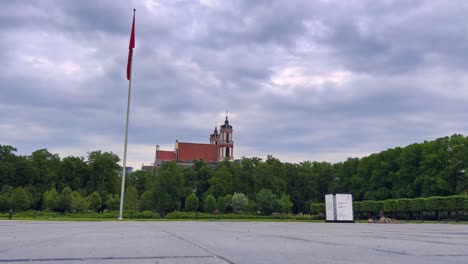 Lithuanian-flag-hanging-on-flagpole-with-church-in-background-at-Lukiskes-square-in-Vilnius,-Lithuania