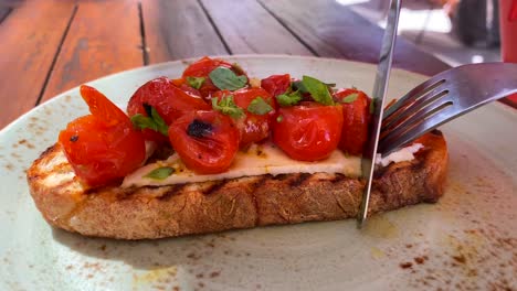 Cutting-a-delicious-bruschetta-sourdough-toast-with-cherry-tomatoes-and-fresh-basil,-traditional-italian-breakfast-brunch-dish-at-a-restaurant,-4K-shot