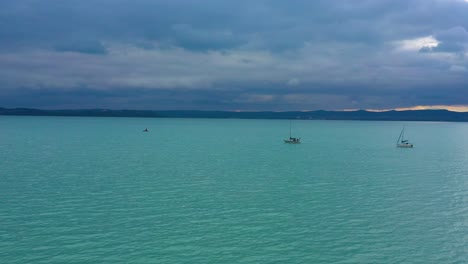Dos-Barcos-Navegando-En-El-Lago-Balaton-Al-Atardecer-Con-Nubes-En-Tiempo-Lluvioso,-Imágenes-De-Drones