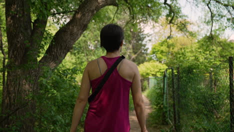 Woman-wearing-pink-tanktop-walking-through-a-forest-trail-next-to-a-fence-on-a-sunny-summer-day