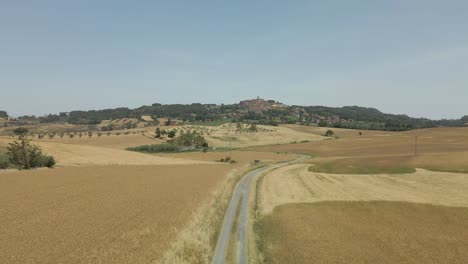 Luftbilder-Der-Toskana-In-Italien-Bebaute-Felder-Sommer,-Flug-über-Eine-Unbefestigte-Straße-Mit-Goldgelben-Kornfeldern-Wunderschöne-Kleine-Bergstadt-Im-Hintergrund