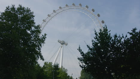 World-Famous-London-Eye-Ferris-Wheel-Landmark-Seen-From-Low-Pedestrian-Perspective-Behind-Trees-With-Red-Double-Deck-Bus-Driving-Through