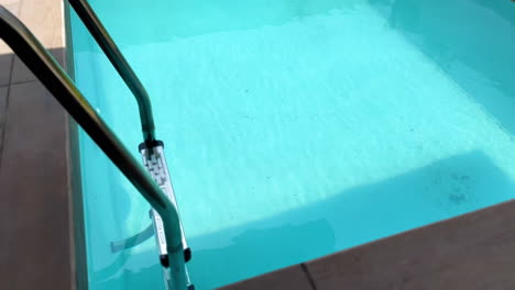 Crystal-clear-turquoise-blue-water-in-a-swimming-pool-with-ladder,-slow-moving-high-angle-shot