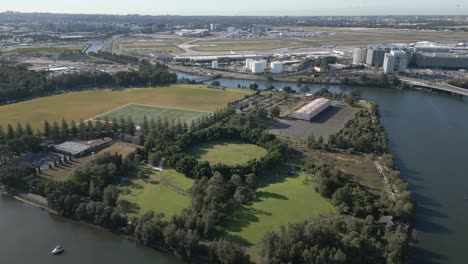 Aerial-zoom-in-flying-over-the-cooks-river,-park-with-green-trees,-playground-and-indoor-and-outdoor-sport-stadium-with-Sydney-Airport-in-the-background