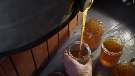 Traditional-Honey-Production-and-Harvest-in-Bee-Farm,-Freshly-Extracted-Golden-Pure-Liquid-Honey-Flowing-from-Extractor-Tank-into-Glass-Jars,-Hand-Holding-the-Jar