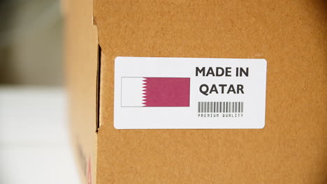 Manos-Aplicando-La-Etiqueta-De-La-Bandera-De-Qatar-En-Una-Caja-De-Cartón-De-Envío-Con-Productos