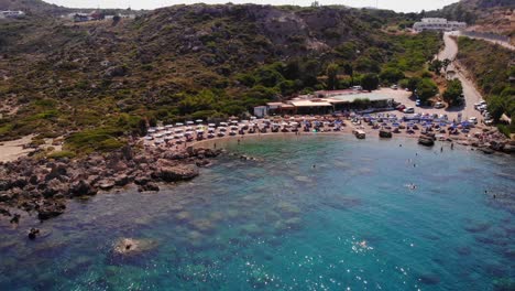 wide-aerial-view-of-Anthony-Quinn-Bay-with-its-azure-blue-waters-and-a-great-beach-full-of-umbrellas-and-sun-beds-with-the-bars-and-restaurants-in-the-background