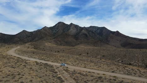 Luftaufnahme-Eines-Weißen-Autos,-Das-An-Einem-Sommertag-Eine-Unbefestigte-Straße-Durch-Die-Wüste-In-Rachel,-Nevada-Hinunterfährt,-Mit-Einem-Berg-Im-Hintergrund-Mit-Blauem-Himmel-Und-Weißen-Wolkenflecken