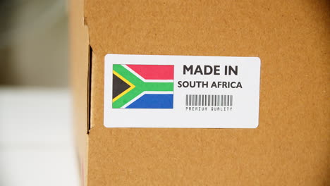 Manos-Aplicando-Etiqueta-De-Bandera-Hecha-En-Sudáfrica-En-Una-Caja-De-Cartón-De-Envío-Con-Productos