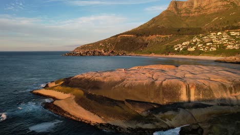 two-girls-walking-on-large-boulders-at-Llandudno-Beach-with-Table-Mountain-behind-in-Cape-Town-at-golden-hour-sunset,-aerial