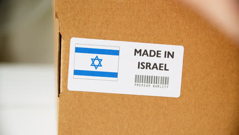 Manos-Aplicando-Etiqueta-De-Bandera-Hecha-En-Israel-En-Una-Caja-De-Cartón-De-Envío-Con-Productos