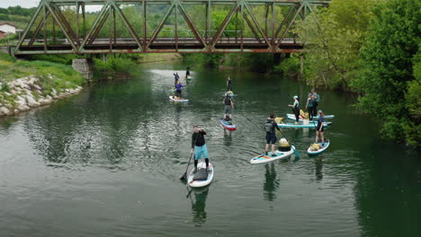 Drone-shot-of-people-paddle-boarding-on-the-river-with-an-iron-bridge-in-the-background