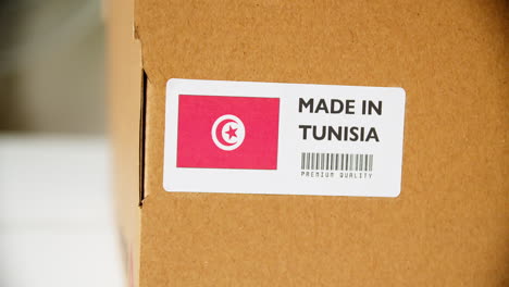 Hands-applying-MADE-IN-TUNISIA-flag-label-on-a-shipping-cardboard-box-with-products