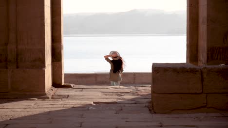 Caucasian-woman-with-hat-running-through-the-ruins-of-Temple-of-Philae-with-lake-in-background-in-Aswan,-Egypt
