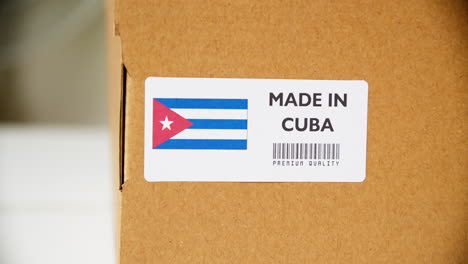 Manos-Aplicando-La-Etiqueta-De-La-Bandera-Hecha-En-Cuba-En-Una-Caja-De-Cartón-De-Envío-Con-Productos