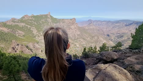 Unrecognizable-blonde-girl-observes-the-view-and-the-Roque-Nublo-from-the-top-of-a-mountain-with-her-back-to-the-viewer