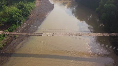 Sliding-drone-shot-of-suspension-bridge-over-the-river-with-motorcycle-crossing-on-it-in-the-morning-with-sun-ray,-central-java,-Indonesia