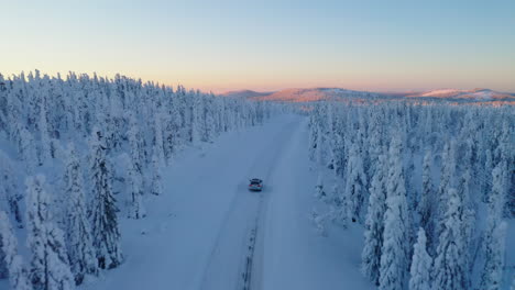 Aerial-view-above-vehicle-travelling-freezing-snowy-remote-woodland-road-towards-sunrise-mountain-landscape