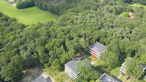 Aerial-of-small-office-buildings-with-solar-panels-on-rooftop-in-a-green-forest-and-a-parking-lot-providing-shadow-to-cars-with-solar-panels