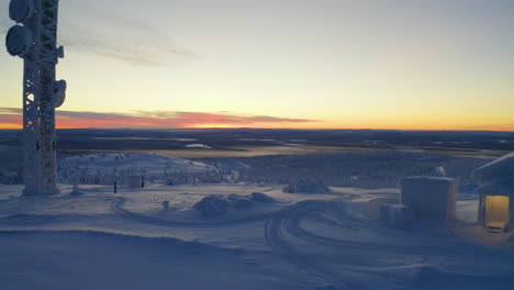 Umgekehrte-Luftaufnahme-Durch-Schneebedeckte-Kabine-Und-Turm-Auf-Dem-Winterlichen-Skandinavischen-Hügelgipfel-Bei-Sonnenaufgang