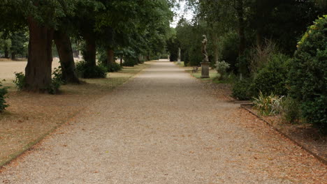 Un-Camino-De-Grava-En-Los-Jardines-De-La-Mansión-Del-Palacio-Con-Estatuas-Y-árboles