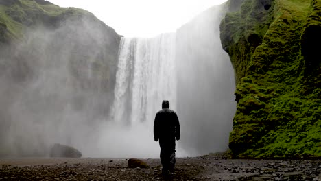 Skogafoss-Wasserfälle-In-Island-Mit-Mann-In-Regenjacke,-Der-In-Zeitlupe-Auf-Die-Wasserfälle-Zugeht