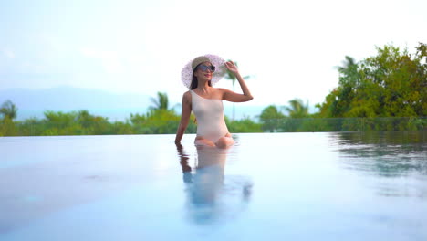 Classy-Slender-Female-in-Swimsuit-and-Summer-Hat-at-Edge-of-Infinity-Pool-of-Tropical-Resort-With-Sea-Horizon-in-Background,-Slow-Motion