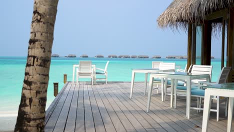 Weiße-Liegestühle-Im-Maldives-Resort-Restaurant-Am-Wasser-Mit-überwasserbungalows-Im-Hintergrund