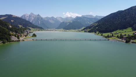 Aerial-shot-of-cars-moving-on-bridge-over-lake-Sihlsee-with-mountain-range-behind