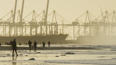 People-silhouettes-walk-on-beach-in-sunset-with-industrial-port-crane-backdrop